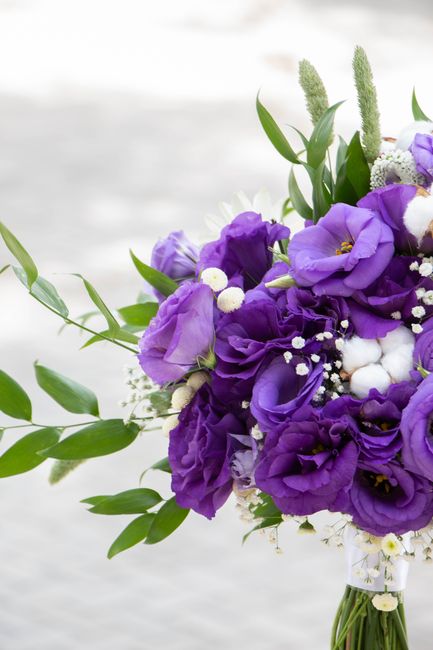 A close-up of a floral bouquet featuring vibrant purple lisianthus and eustoma flowers, complemented by delicate white baby's breath and soft cotton blooms, all arranged with lush green leaves and lon