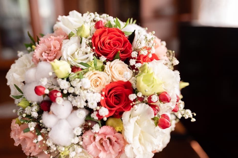 A close-up view of a fresh flower bouquet arranged with red roses, pale peach roses, white lisianthus, pink and white carnations, sprigs of baby's breath, and small red hypericum berries. The bouquet