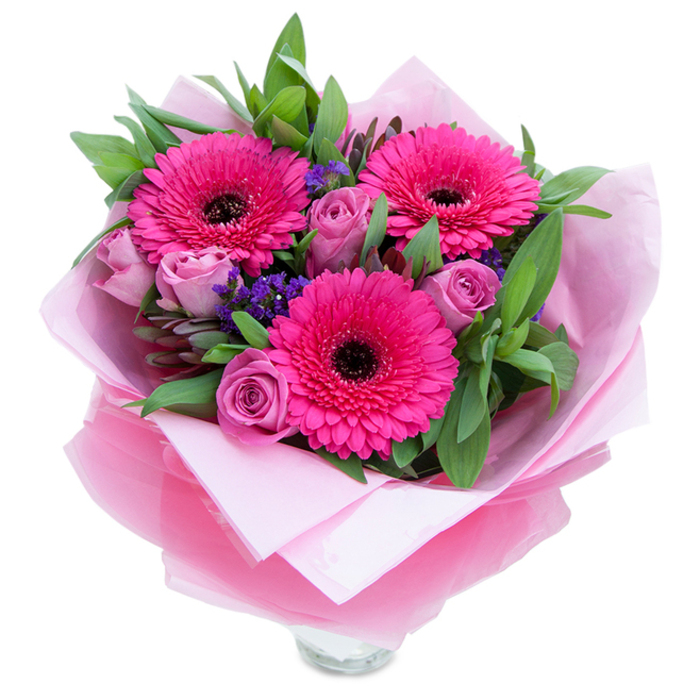 The charm of this handtied bouquet lies in the balance of playfulness and grace: three fuchsia Gerbera daisies, each with numerous petal layers and a deep, nearly-black centre, open boldly amid a softer chorus of lavender-pink roses whose tightly wrapped petals hint at unfolding moments. Tiny bunches of purple statice punctuate the arrangement like small wildflowers picked from a verge, adding cool contrast and a papery texture that complements the velvety rose petals. Surrounding leaves-broad and glossy, reminiscent of salal, with the occasional eucalyptus stem-provide a rich green framework that feels freshly gathered from a garden bed. The bouquet is swathed in a pale-pink matte tissue layer and finished with an outer sheet of slightly crinkled, translucent fuchsia-pink cellophane; together they make the presentation feel intimate and celebratory, the cellophane catching light and the tissue softening the palette. The base shows the top rim of a clear glass vase, indicating the flowers are prepared for instant display and enjoyment. Scent is gentle here: a soft rose perfume tempered by green, watery notes from the foliage. I picture this arrangement placed on a hallway table in a Crayford home or offered as a thoughtful gift after an afternoon visiting Hall Place gardens, its colours designed to lift spirits and mark a moment-be it a birthday, a quiet anniversary, or a spontaneous 'you matter' delivery. Each element reflects careful floral craft: considered colour layering, textural contrast, and an eye for hospitality that suits local Crayford settings and celebrations.